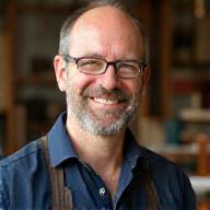 Portræt af Lars Pedersen, en erfaren restaurator med et varmt smil, der holder et stykke antikt træmøbel på et værksted med historiske værktøjer.